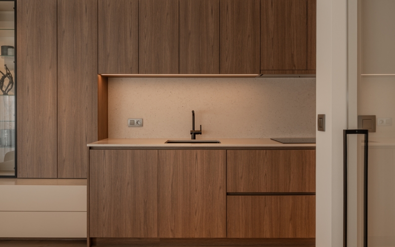 Moderne keuken met ingebouwde houten kasten en lades, een licht aanrechtblad, zwarte kraan en verlichting onder de bovenkasten.
