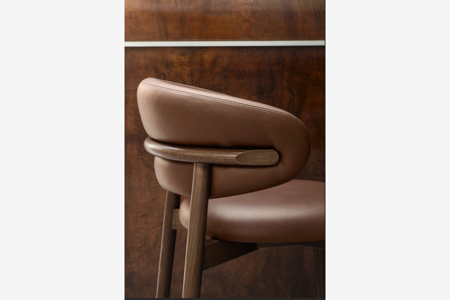 Bruinleren stoel met houten rugleuning en donkere houten poten, met een houten muur op de achtergrond.