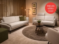 Woonkamer met grote beige bank, kleinere beige bank, ronde salontafel en groene