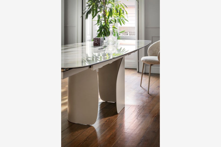 Bovenkant en poten van een moderne beige eettafel met glazen blad, met een stoel op de achtergrond.