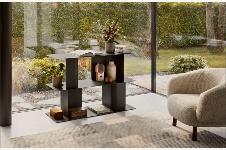 Zwart metalen lage boekenkast met vakken, met decoratie en boeken, naast een lichte fauteuil bij een raam.